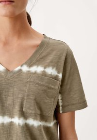 Olivově zelené tričko s výstřihem do V a bílým tie-dye vzorem, vybavené přední kapsou a krátkými rukávy, vyrobené z texturovaného materiálu.