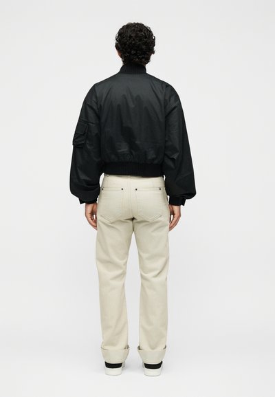 Άτομο με σγουρά μαλλιά που φοράει μαύρο bomber μπουφάν και μπεζ παντελόνια που είναι τυλιγμένα προς τα πάνω, στέκεται αντικρίζοντας ένα απλό λευκό φόντο.