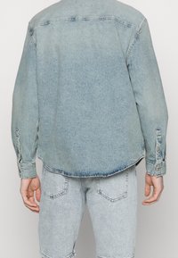 Camicia di denim azzurro chiaro con colletto appuntito, maniche lunghe e polsini con bottoni. Caratterizzata da un orlo curvo e una texture sbiadita.