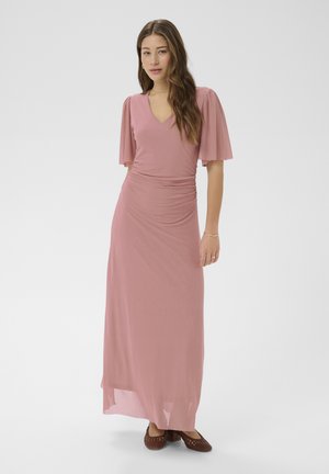 Žena v dlhých ružovkastých maxi šatách s výstrihom do V a nahými rukávmi a hnedými pletenými nízkymi topánkami, stojaca pred jednoduchým pozadím.
