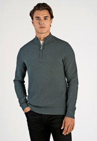 Dunkeltürkisfarbener Pullover mit einem Viertelreißverschluss und strukturiertem Strickdesign. Gerippte Bündchen und Saum, getragen über einem weißen Hemd und schwarzen Hosen.