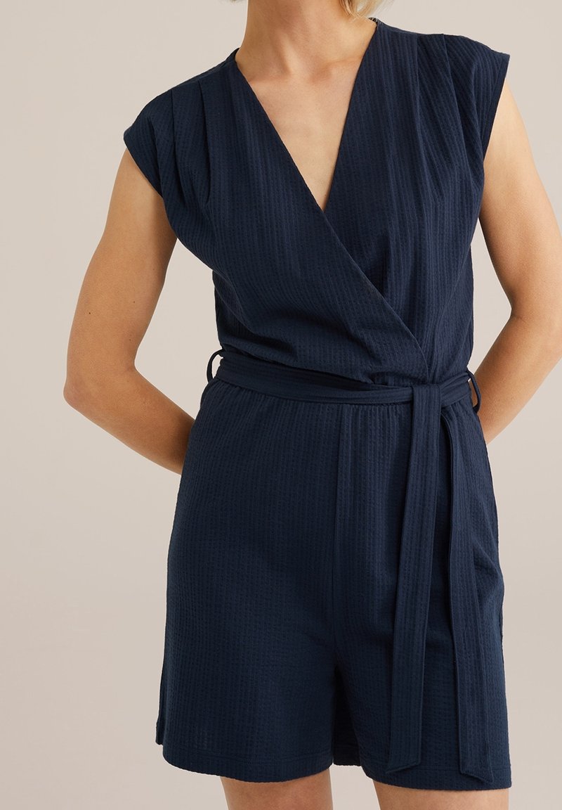 Femme portant une combinaison courte sans manches bleu marine texturée avec un profond décolleté en V et une ceinture nouée à la taille, debout devant un fond uni.