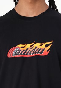 Juodas medvilninis marškinėlis su ryškiu raudonai oranžiniu ugnies dizainu ir žodžiu "cadidas" stilizuotu šriftu per priekinę dalį.
