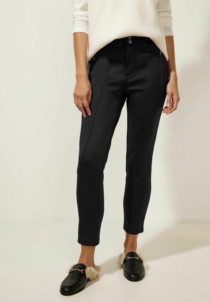 Femme portant un pantalon noir ajusté, un pull blanc côtelé et des mocassins en cuir noir à enfiler doublés de fourrure, debout sur un sol blanc.