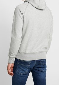 Sudadera gris con capucha y textura suave, mangas raglán y dobladillo de canalé, combinada con jeans de denim azul oscuro con bolsillos traseros.