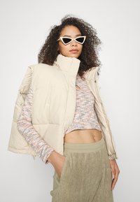 Colete puffer bege com colarinho alto, blusa de malha texturizada em tons multicoloridos, calças de veludo cotelê verde-azeitona e óculos de sol brancos em formato cat-eye.