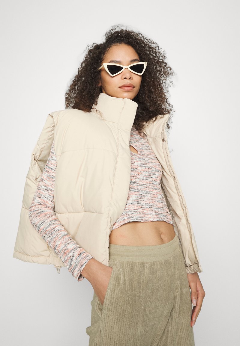 Colete puffer bege com colarinho alto, blusa de malha texturizada em tons multicoloridos, calças de veludo cotelê verde-azeitona e óculos de sol brancos em formato cat-eye.