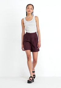 Stripped tank top, maroon pleated shorts, and chunky black sandals. Features include a button closure and hoop earrings, standing against a white backdrop.