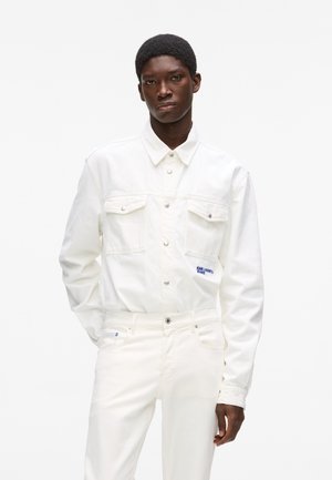 Bijela traper jakna s dva prednja džepa, zatvaranjem na kopče i plavim logotipom kao naglaskom. Uparena s bijelim trapericama uskog kroja.