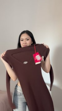 Fiatal nő hosszú ujjú barna ruhát tart fel egy piros árjelzéssel, egyszerű világos fal háttér előtt.