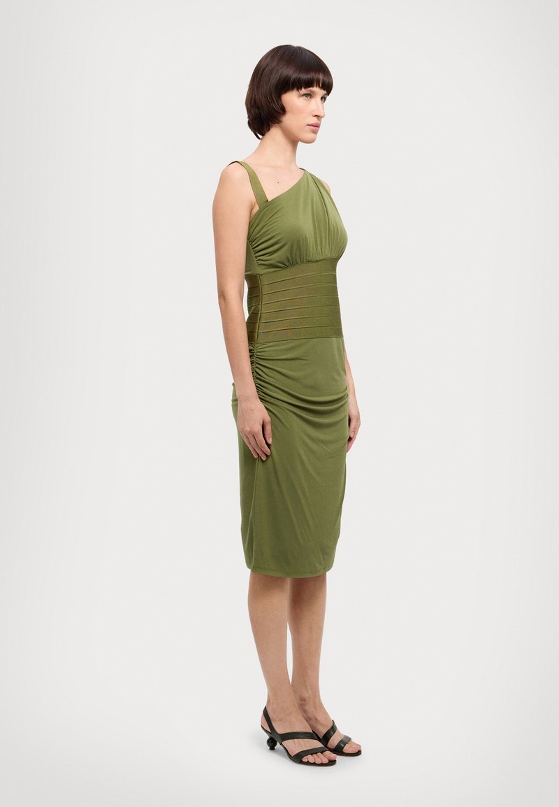 Robe verte à une épaule avec des détails froncés et un corsage ajusté. Le matériau est lisse avec une texture extensible. Sandales avec des brides noires.