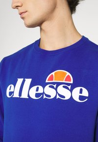 Sweat-shirt bleu avec un logo blanc surmonté d'un arc orange et rouge au-dessus du texte « ellesse ». Col rond et texture douce.