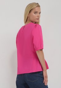 Donna con capelli biondi che indossa una blusa rosa brillante con maniche a lunghezza gomito e jeans blu, che guarda oltre la spalla.