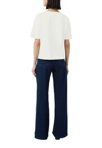 Weißes kürzesärmeliges Oberteil, kombiniert mit hochgeschnittenen, dunkelblauen, weit geschnittenen Jeans. Der Stoff wirkt weich und hat ein klares Design ohne sichtbare Muster.