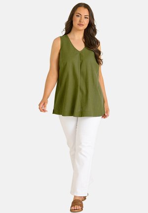 Femme portant une tunique olive sans manches à col en V, un pantalon blanc et des sandales marron ouvertes, marchant vers l'avant sur un fond clair.