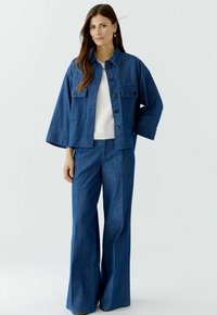 Denimoutfit met een cropped jack met grote zakken en wijde broekspijpen. De kleur is een medium blauw met een gladde textuur.