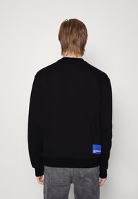 Felpa nera con maniche lunghe e polsini a coste. Presenta un'etichetta blu sull'orlo sinistro posteriore. Il materiale appare morbido e informale.
