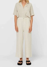 Donna che indossa una camicia beige a maniche corte con bottoni e pantaloni larghi abbinati, con sandali con piattaforma nera su uno sfondo neutro.