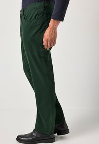Man met donkere groene broek, zwarte nette schoenen en een langemouwen donker shirt, staand tegen een effen lichte achtergrond.