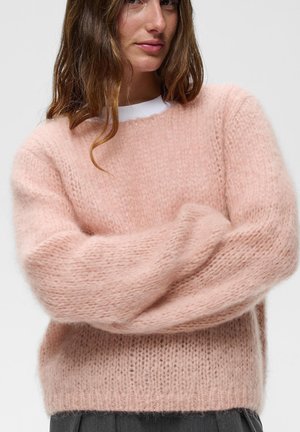 Kvinde med langt brunt hår iført en blød, fluffy lyserød strikket sweater, der krydser armene foran en ensfarvet lys baggrund.