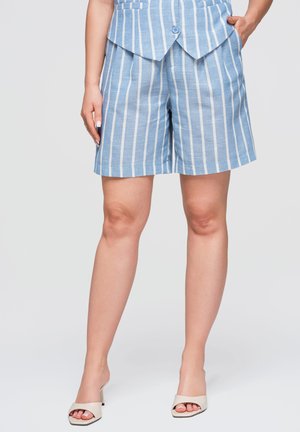 Femme portant un short rayé vertical bleu et blanc assorti à un haut coordonné et des sandales blanches à talons ouverts, debout avec une main dans la poche.