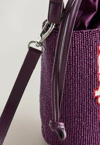 Gros plan sur un sac à main à perles violettes avec une bandoulière en cuir et une boucle argentée fixée à un panneau latéral texturé.