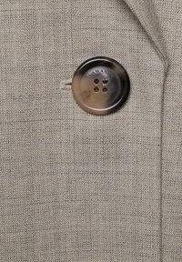 Prim-plan al unui nasture de plastic maro cusut pe o țesătură texturată bej, evidențiind o găurică cusută pentru nasture și detalii subtile ale țesăturii.