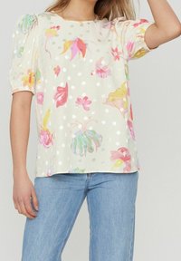 Kvinna i krämfärgad blus med flerfärgad blommig och prickig design samt puffärmar, kombinerad med ljusblåjeans.