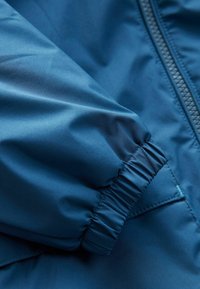 Blauwe, lichtgewicht jasstof met een gladde textuur; beschikt over elastische manchetten en een rits aan de zijkant.