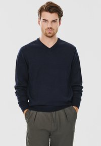 Granatowy sweter w serek wykonany z miękkiego materiału, z długimi rękawami i ribbowymi mankietami. Noszony z szarymi spodniami o luźnym kroju.