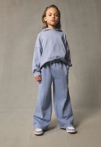 Hoodie bleu clair avec épaules tombantes, associé à un pantalon de survêtement ample assorti. Baskets blanches avec des accents gris. Tissu simple et doux.