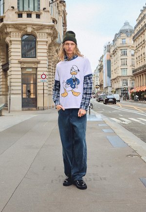 Biała koszulka z kolorową grafiką Kaczora Donalda, nałożona na niebieską koszulę w kratę, w zestawie z ciemnymi dżinsami i czarnymi butami.