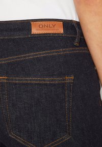 Pantalones vaqueros de mezclilla oscura con costuras en color naranja, que presentan una etiqueta de cuero rectangular etiquetada "ONLY". El diseño incluye dos bolsillos traseros.