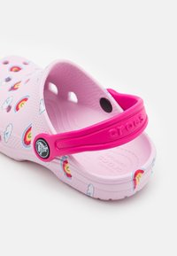 Różowe klapki Crocs ozdobione tęczą i chmurkami, z regulowanym jasnoróżowym paskiem na pięcie, na białym tle.