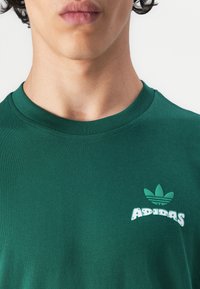 Zöld pamut póló kerek nyakkal, fehér és zöld Adidas logóval a mellkason, sima, egyszínű textúrával.