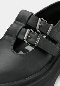 Fekete bőrcipő lekerekített orral, kivágott mintával és két ezüst csattal ellátott szíjjal. Sima textúra, vastag talppal.