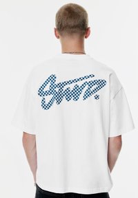 Hvid bomulds T-shirt med løs pasform, der har et blåt ternet grafisk design på ryggen. Korte ærmer, rund hals.