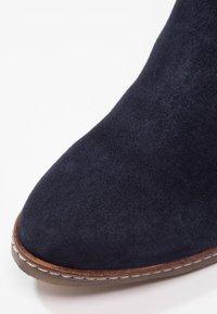 Gros plan sur l'avant d'une chaussure en daim bleu marine avec une couture de renfort marron et un bord de semelle en caoutchouc.
