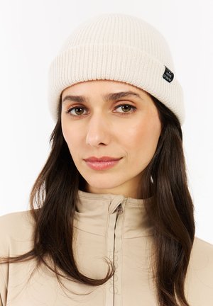 Witte geribbelde gebreide beanie met een snug fit, voorzien van een klein zwart label. De achtergrond is eenvoudig en richt de aandacht op de textuur en stijl van de muts.
