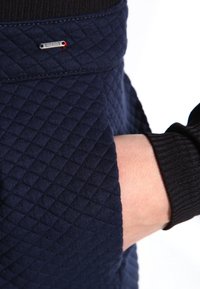 Marint quiltat tyg med ett strukturerat diamantmönster, med en liten silverdetalj nära fickan. Svarta ribbade manschetter.