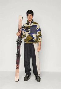 Fleece jacket with multicolored camouflage pattern, zip pocket, and black pants with beige accents. Holding a ski with bindings.