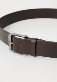 Barna bőr öv ezüst színű fém csattal. Simább textúrával és több lyukkal a beállításhoz. A bőrre nyomtatott logóval.
