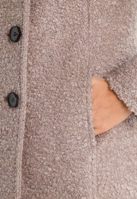 Beige tekstureret uldfrakke med sorte knapper og en hånd placeret inde i lommen på højre side.