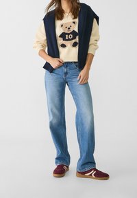 Pull crème avec un motif d'ours tricoté, associé à un jean évasé bleu clair et des baskets bordeaux avec des accents blancs, le tout superposé d'une veste bleu marine.