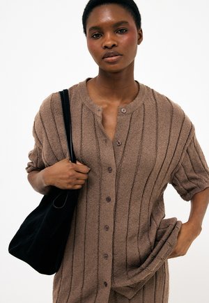 Kobieta w brązowej, prążkowanej, zapinanej na guziki bluzce z dzianiny, trzymająca czarną torebkę na ramię jedną ręką, a drugą dotykająca swojego boku.