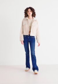 Beige cropped jacket with two front pockets and button closure, paired with dark blue flared jeans and white sneakers. Smooth texture.