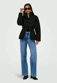 Donna con giacca nera trapuntata, jeans a vita alta blu, stivali neri, grandi orecchini a cerchio e occhiali da sole scuri che aggiusta la montatura.