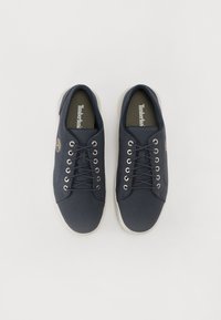 Tumši jūras zila auduma sporta apavi ar baltām gumijas zēmēm, ar metāla acu caurumiem un auklām, kā arī Timberland logo sānos.