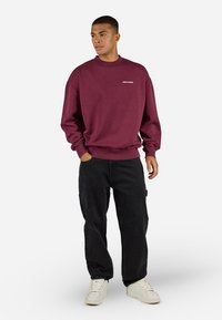 Maroon sweatshirt with ribbed cuffs and neckline, featuring a white logo. Paired with relaxed-fit black pants and white sneakers.