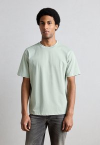 Svetlozelené bavlnené tričko s krátkymi rukávmi, okrúhlym výstrihom, voľného strihu, doplnené tmavosivými džínsami a jednoduchým pozadím.